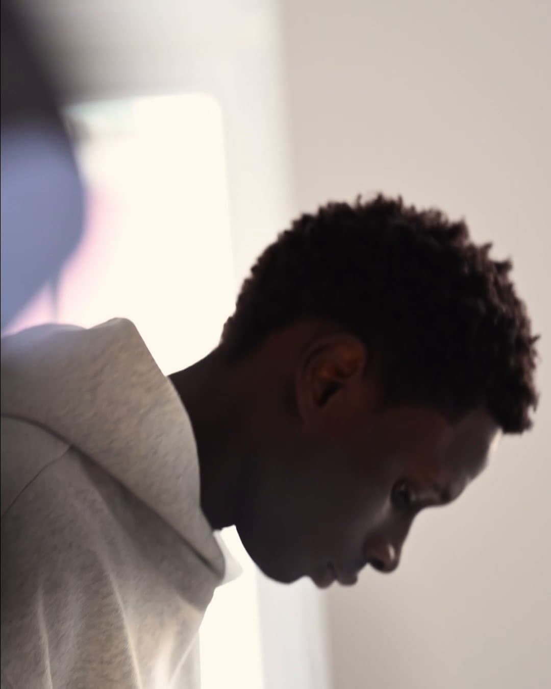 Model in a grey hoodie in profile, looking down, in soft window light