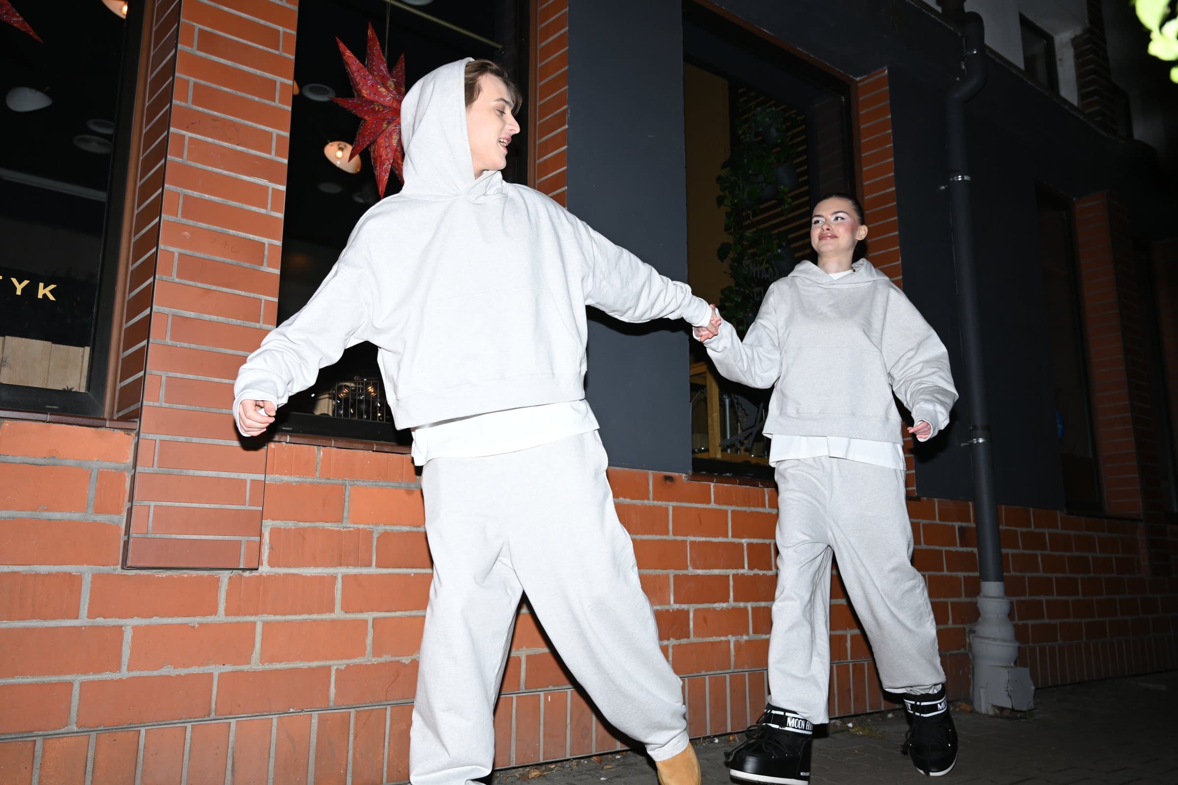 Two models in grey tracksuits stepping out along a brick wall at night, lit by flash