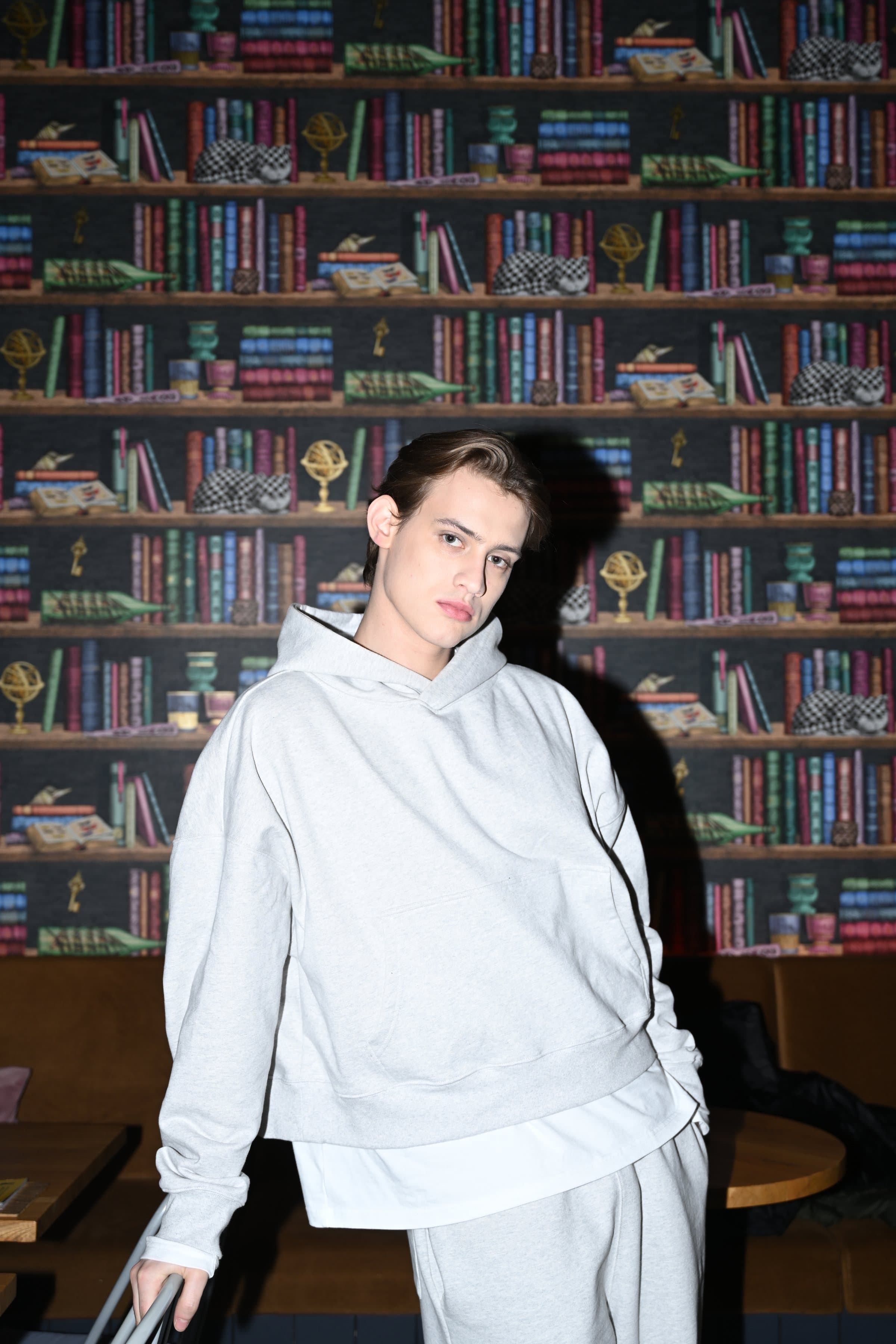Model in a grey hoodie leaning against a wall of bookshelves