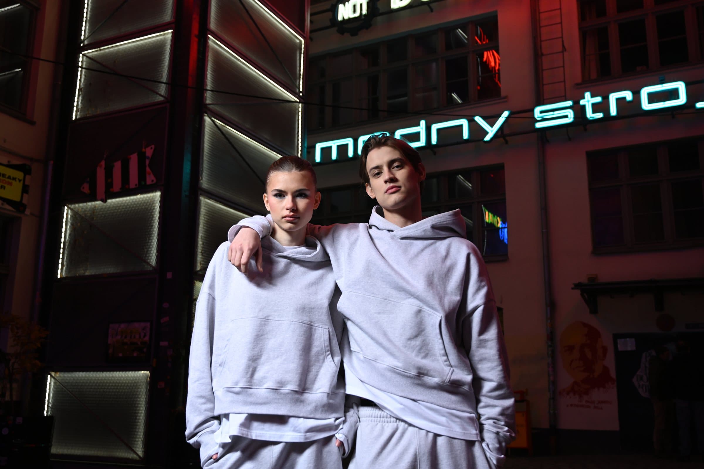 Two models in grey hoodies posing close together under a green neon sign at night