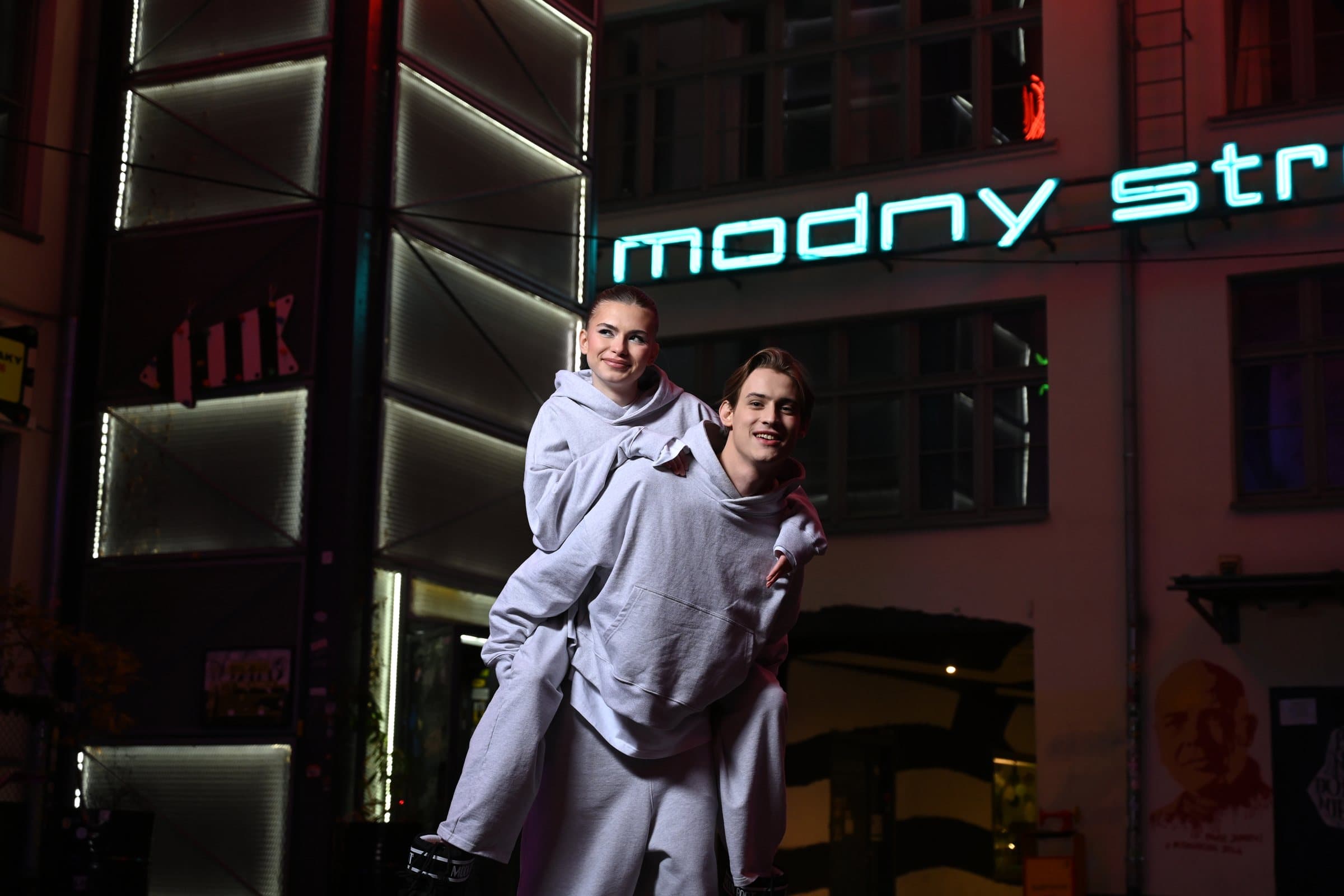 Two models in grey tracksuits posing under a large neon sign in a narrow street