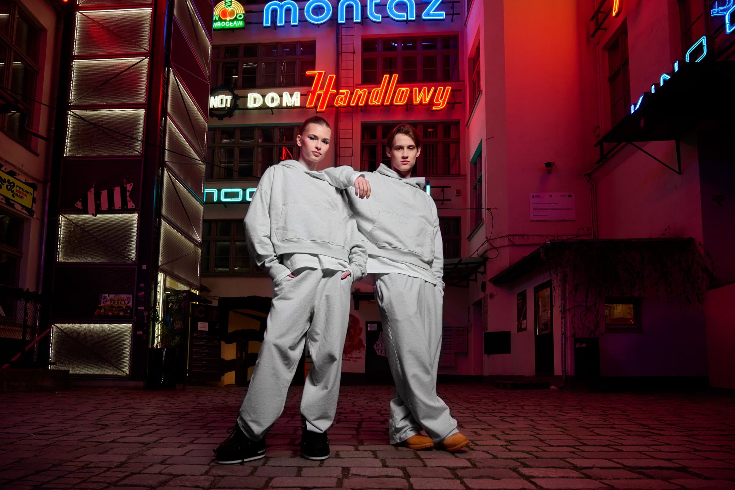 Two models in light grey loungewear standing in a neon-lit street at night