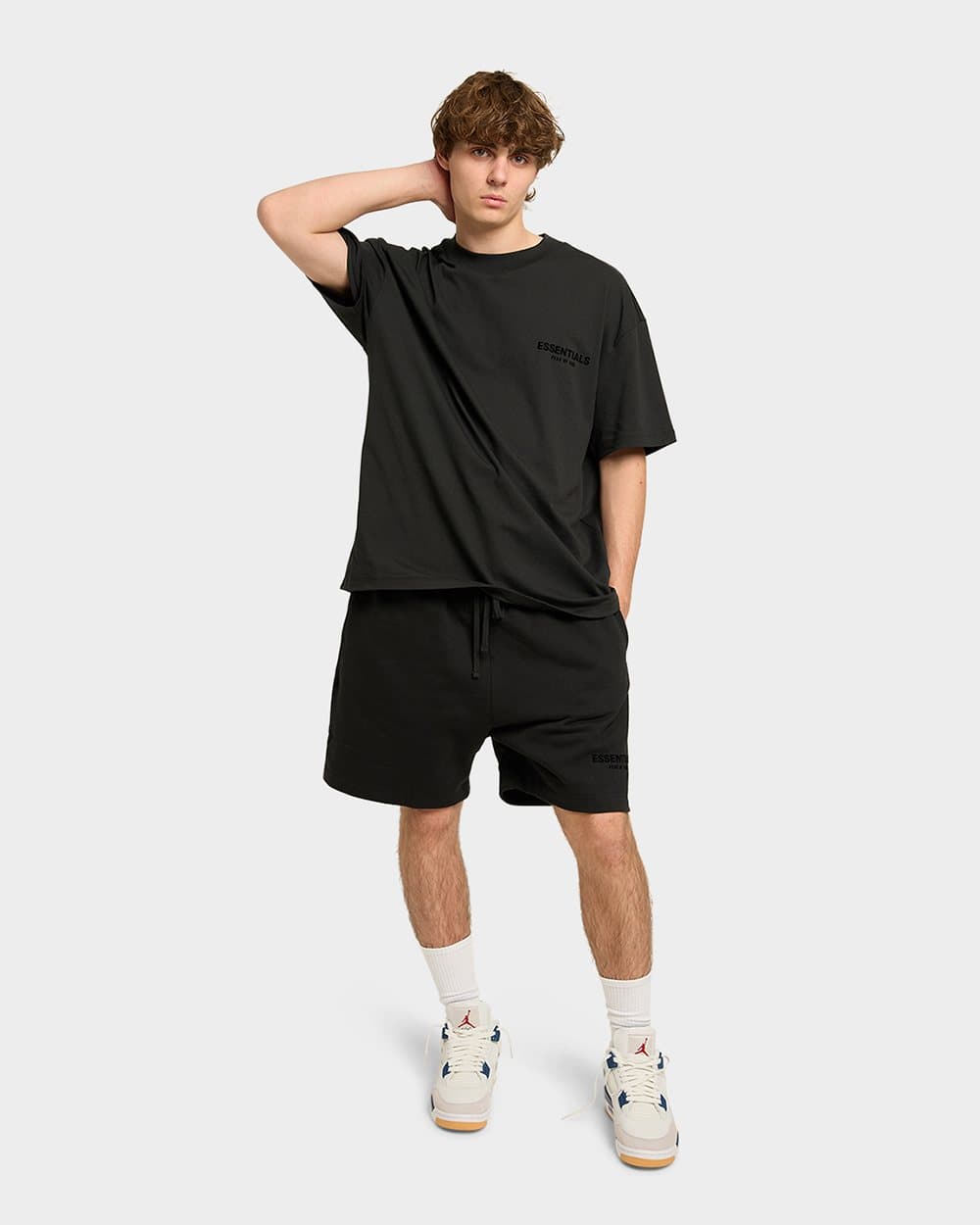Model in a black T-shirt and shorts with one hand behind his head on a light backdrop