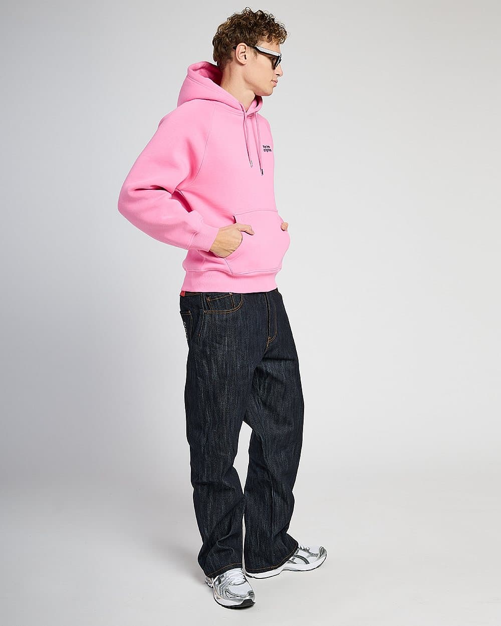 Model in a pink hoodie, sunglasses and dark jeans on a grey studio backdrop