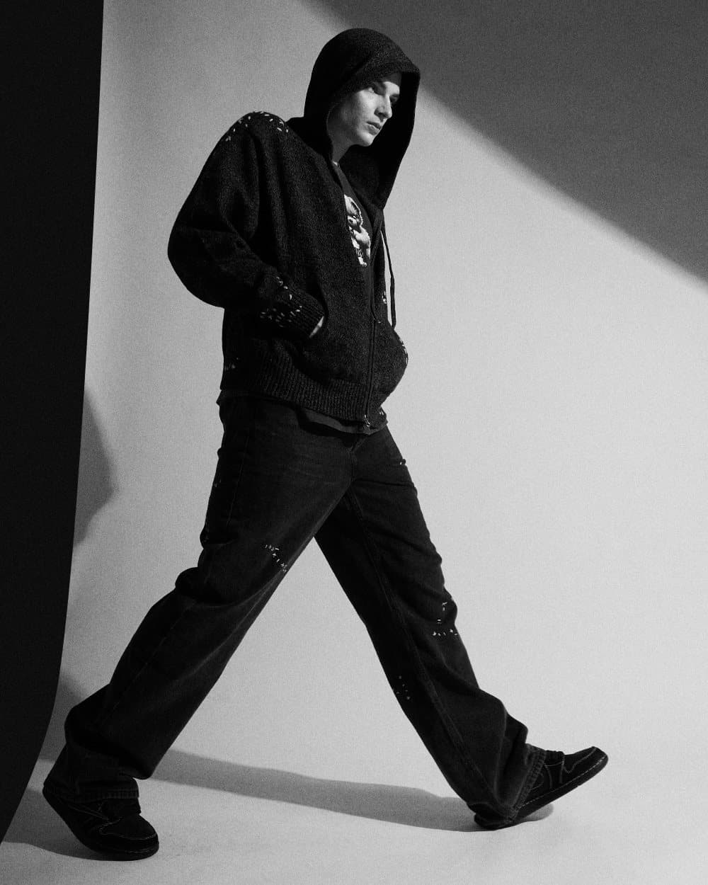 Model in a black hooded jacket striding across a studio set with strong shadow