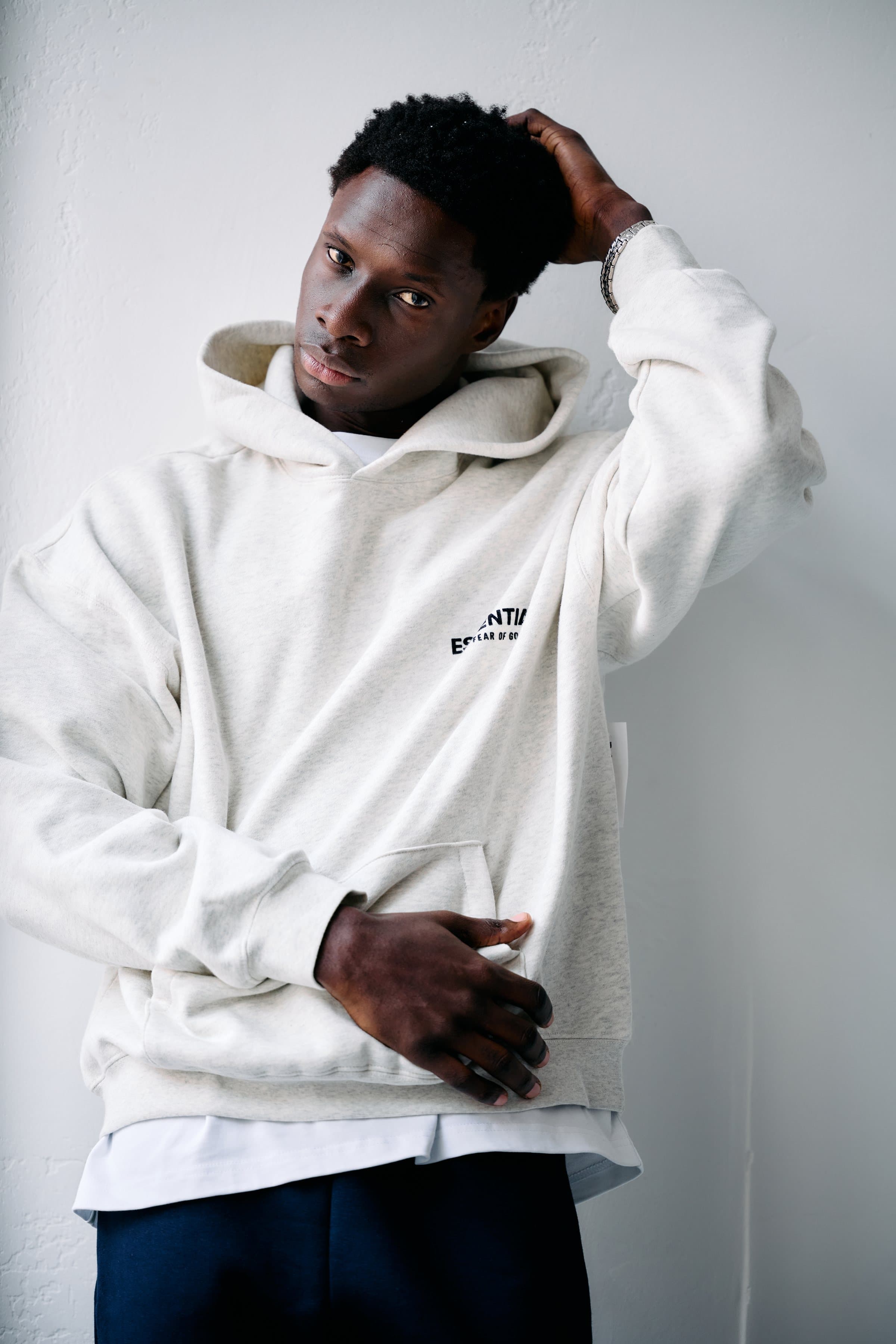 Model in an oatmeal hoodie leaning on a white wall with one hand in his hair