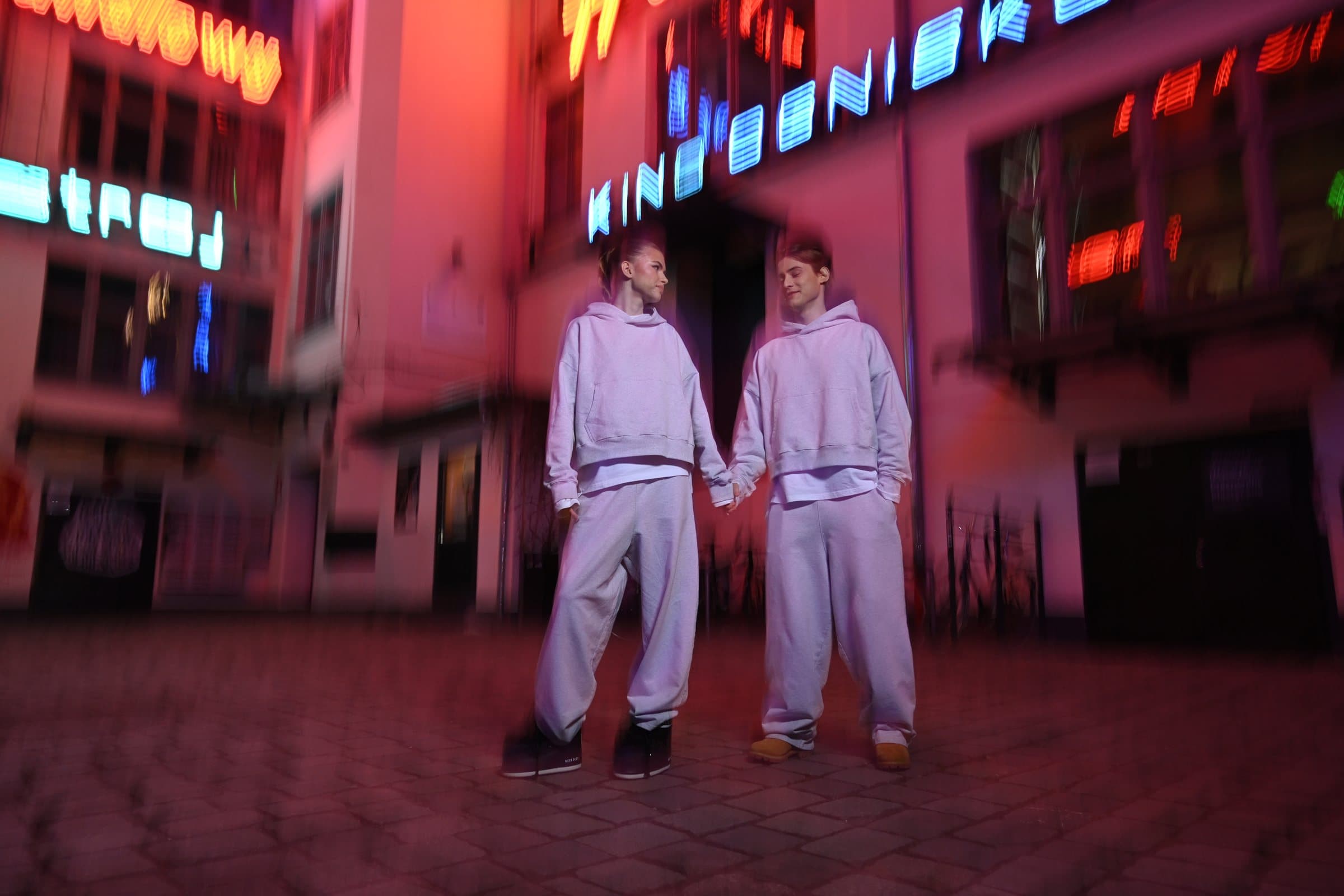 Two models in grey tracksuits on a cobbled street under pink and red neon signs at night