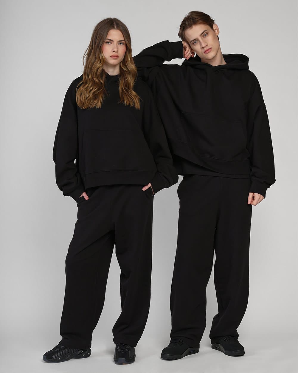 Two models in black sweatshirts and trousers posing together on a grey studio backdrop