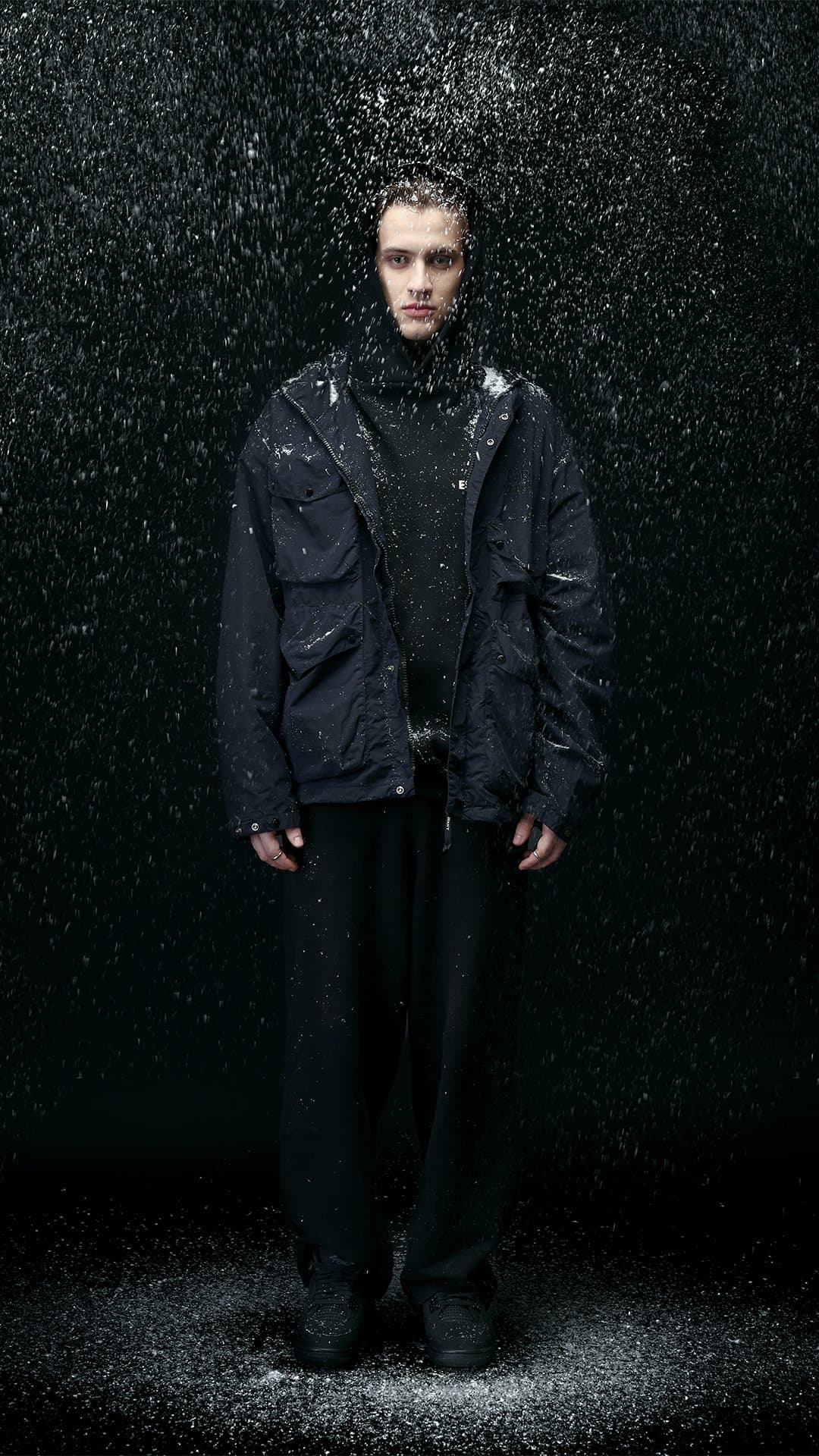 Model in a dark technical jacket in a dark set with falling white particles